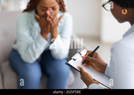Betonte afroamerikanische Frau, die Psychologin konsultiert, weint im medizinischen Büro, selektiver Fokus. Psychiater hilft verzweifelte schwarze Dame mit ner Stockfoto