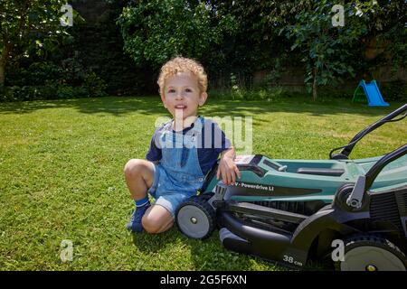 Ein weißer kaukasischer kleiner Junge im Alter von fast 3 Jahren mit lockigen blonden Haaren, die in Denim-Latzhose gekleidet sind, kniet an einem sonnigen Tag in England bei einem Rasenmäher im Garten Stockfoto