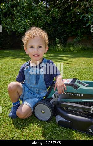 Ein weißer kaukasischer kleiner Junge im Alter von fast 3 Jahren mit lockigen blonden Haaren, die in Denim-Latzhose gekleidet sind, kniet an einem sonnigen Tag in England bei einem Rasenmäher im Garten Stockfoto