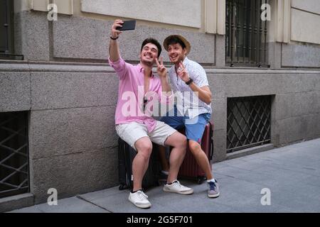 Schwule Touristen, die ein Selfie machen, lachen und das Siegeszeichen auf ihren Koffern machen. Stockfoto