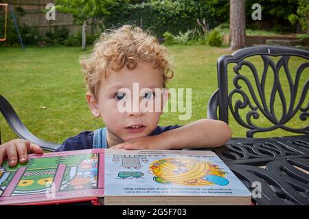 Ein weißer kaukasischer kleiner Junge mit lockig blondem Haar im Alter von 2 -3 Jahren sitzt an einem sonnigen Tag in England mit einem Kinderbuch vor der Kamera an einem Tisch in einem Garten Stockfoto