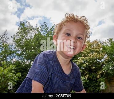 Ein lächelnder, fröhlicher weißer kaukasischer kleiner Junge im Alter von fast 3 Jahren mit lockigen blonden Haaren in einem Garten, der ein blaues Oberteil trägt, lächelt an einem sonnigen Tag in en an der Kamera Stockfoto