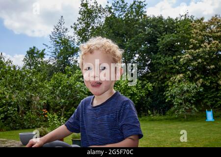 Ein glücklicher weißer kaukasischer kleiner Junge im Alter von fast 3 Jahren mit lockigen blonden Haaren in einem Garten mit blauem Oberteil sticht seine Zunge an einem sonnigen Tag in England aus Stockfoto