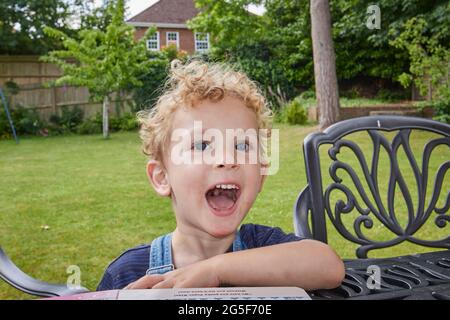 Ein glücklicher weißer kaukasischer kleiner Junge im Alter von fast 3 Jahren mit lockig blonden Haaren sitzt an einem Tisch in einem Garten an einem sonnigen Tag in England mit offenem Mund Stockfoto
