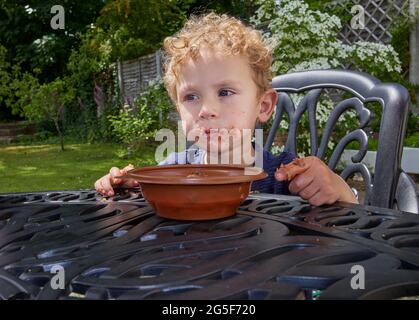 Ein klebriger weißer kaukasischer kleiner Junge im Alter von fast 3 Jahren mit lockigem blondem Haar sitzt im Freien mit einem schmuddeligen braunen Mund, der Schokoladenkuchen isst Stockfoto