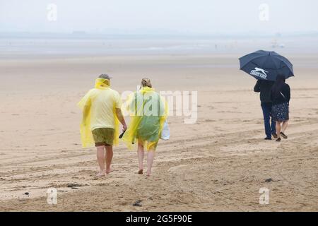 Camber, East Sussex, Großbritannien. 27 Juni 2021. Wetter in Großbritannien: Das später erwartete Regenwetter ist früh an den Sandstränden von Camber Sands in East Sussex angekommen. Einige Familien wollen den Tag genießen, wenn sie mit Regenschirmen und Regenponchos ankommen. Foto-Kredit: Paul Lawrenson /Alamy Live Nachrichten Stockfoto