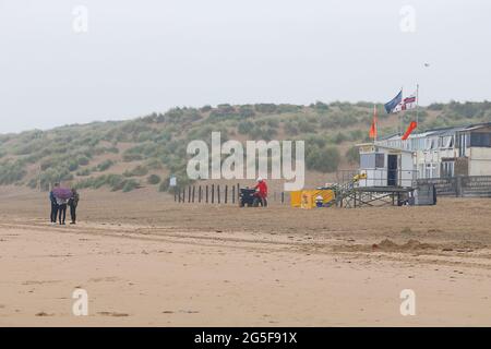 Camber, East Sussex, Großbritannien. 27 Juni 2021. Wetter in Großbritannien: Das später erwartete Regenwetter ist früh an den Sandstränden von Camber Sands in East Sussex angekommen. Einige Familien wollen den Tag genießen, wenn sie mit Regenschirmen und Regenponchos ankommen. Foto-Kredit: Paul Lawrenson /Alamy Live Nachrichten Stockfoto