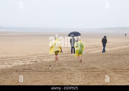 Camber, East Sussex, Großbritannien. 27 Juni 2021. Wetter in Großbritannien: Das später erwartete Regenwetter ist früh an den Sandstränden von Camber Sands in East Sussex angekommen. Einige Familien wollen den Tag genießen, wenn sie mit Regenschirmen und Regenponchos ankommen. Foto-Kredit: Paul Lawrenson /Alamy Live Nachrichten Stockfoto