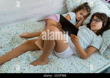 Zwei kaukasische Schwestern, 2 und 9 Jahre alt, liegen mit einem Tablet in ihrem Schlafzimmer auf ihrem Bett. Stockfoto