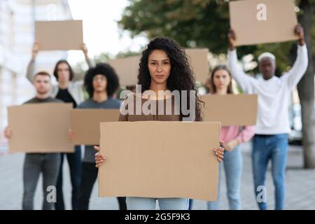 Aktive, hübsche junge Frau, die eine Gruppe von Demonstranten auf der Straße führt. Internationale, vielfältige Gruppe von Menschen mit leeren Plakaten, die gegen Brummen protestierten Stockfoto