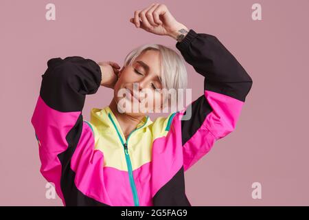 Weißhaarige Frau in einer bunten Jacke, die isoliert auf der Kamera über der lila Wand tanzt Stockfoto