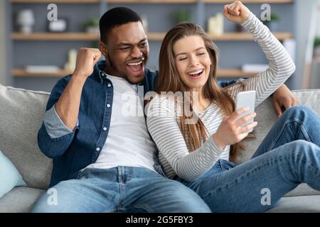 Aufgeregt Interracial Paar Blick Auf Smartphone-Bildschirm Und Ausrufen Mit Freude Beim Entspannen Zusammen Zu Hause, Emotionale Multikulturelle Lovers Stockfoto