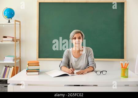 Der Lehrer lehrt. Ernsthafte ältere Lehrerin, die beim Unterricht auf die Kamera schaut, auf dem Hintergrund eines Greenboards im Klassenzimmer. Stockfoto
