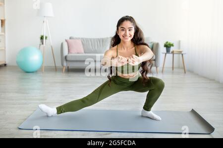 Sperren Sie Ihr Heimtraining ab. Sportliche junge indische Dame, die sich auf einer Yogamatte im Wohnzimmer die Beine streckt, freier Platz. Positive Eastern Frau tut Flexibilität sehr gut Stockfoto