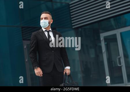 Selbstbewusster Geschäftsmann mittleren Alters, der im Freien vor dem Business Center steht, eine Gesichtsmaske mit schwarzem Anzug und Panorama mit Kopierfläche trägt Stockfoto