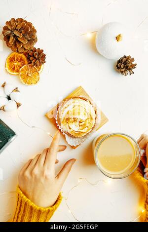 Flache Hände der Frauen berühren Kaffeebecher und Tartlet. Mädchen in Pullover ist mit Frühstück. Süßes Dessert, Getränk, Tannenzapfen und Dekorationen auf weißem Tisch. Hintergrund für festliche Postkarte. Stockfoto
