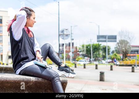 Junge Latina-Frau, die mit einem Notizblock auf einem Bankpark sitzt, ihr Haar berührt und den Horizont betrachtet Stockfoto