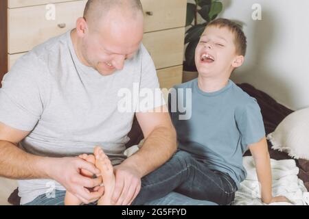 Vater spielt mit seinem Sohn 7-10, der auf dem Boden sitzt. Papa kitzelt die Füße der Kinder. Familie, die Spaß hat Stockfoto