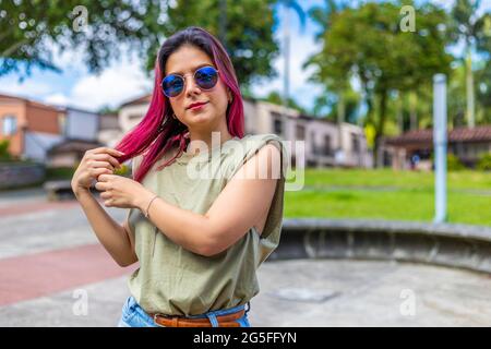 Junge Latina-Frau im Park mit blauer Brille, die ihr fuchsiaes Haar mit ihren Händen arrangiert und die Kamera anschaut Stockfoto