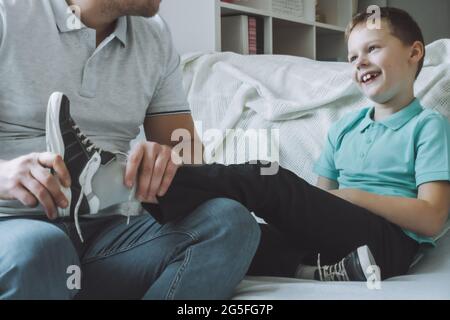 Vater und Sohn sitzen auf der Couch. Vater hilft dem Kind, sich auszuziehen oder Schuhe anzuziehen, lacht Kind Stockfoto