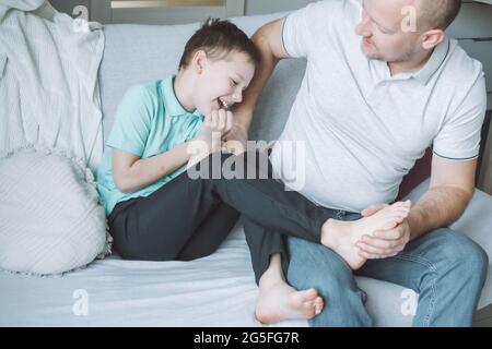 Vater spielt mit seinem Sohn 7-10 auf der Couch sitzend. Papa kitzelt die Füße der Kinder. Familie, die Spaß hat Stockfoto