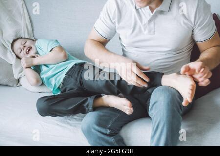 Vater spielt mit seinem Sohn 7-10 auf der Couch sitzend. Papa kitzelt die Füße der Kinder. Familie, die Spaß hat Stockfoto