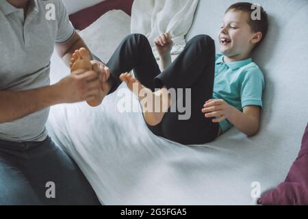 Vater spielt mit seinem Sohn 7-10 auf der Couch sitzend. Papa kitzelt die Füße der Kinder. Familie, die Spaß hat Stockfoto