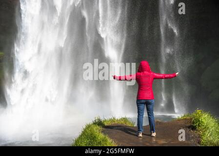 Frau mit ausgestreckten Armen am Fuße eines majestätischen Wasserfalls an einem sonnigen Sommertag. Kraft in der Natur. Stockfoto