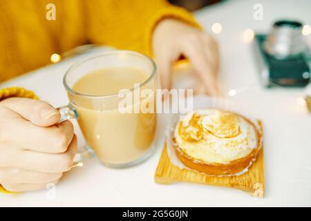 Die Hände der Frauen berühren Kaffeebecher und Tartlet. Mädchen in gestrickten Pullover ist mit Frühstück. Romantische Atmosphäre. Süßes Dessert, erfrischendes Getränk und Fotokamera auf weißem Tisch. Stillleben ästhetisch. Stockfoto