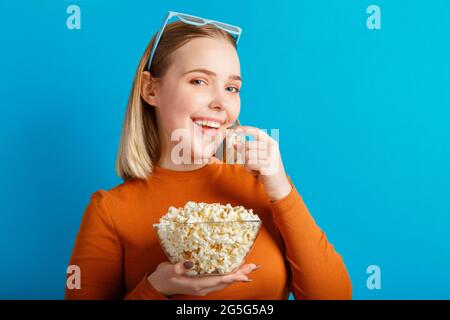 Junge Frau in einer Kinobrille beim Betrachten von 3d-Filmen. Lächelndes Teenager Mädchen Filmbetrachter in Gläsern isst Popcorn mit Kopierraum isoliert über blauer Farbe Stockfoto