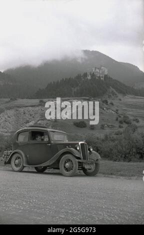 1934, historisch, Touring Bohemia, der westliche Teil der Tschechoslowakei vor WW2. Ein junger Mann, der in seinem Auto sitzt, ein britischer Morris Eight auf einer Landstraße, mit den Sudetenhügelern in der Ferne. Dieses Gebiet der Tschechoslowakei war zu dieser Zeit als Sudetenland bekannt, der historische Name für das sudetendeutsche Volk, das in den westtschechischen Ländern Böhmens und anderen Grenzgebieten lebt. Stockfoto