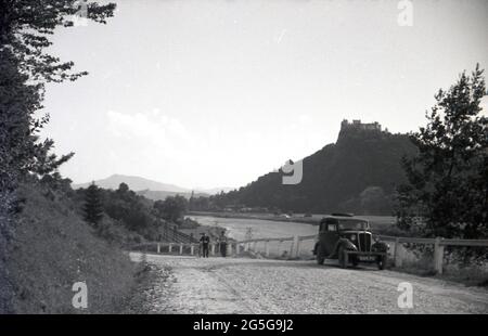 1930s, hisotrical, Touren durch Czechoslavia vor WW2, ein britisches Auto, ein Morris 8, Geparkt auf einer ländlichen Schotterstraße, neben einer Eisenbahnstrecke und einem Fluss, mit einer Burg auf einem Hügel im Wald im Hintergrund. Dieses Gebiet der Tschechoslowakei war zu dieser Zeit als Sudetenland bekannt und wurde auch als Sudetenland bezeichnet, da es sich um viele deutsche Volksgruppen handelte, die in den westtschechischen Ländern Böhmens lebten. Stockfoto