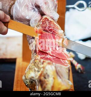 Der Metzger schneidet mit Nahaufnahme des Messers und selektivem Fokus Staus. Männliche Hände des Verkäufers schneiden Scheiben aus Jamon in Fleischgeschäft, Supermarkt Stockfoto