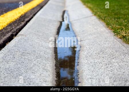 Fließendes Regenwasser im Kanal ist ein Zementgraben eines Drainage-Systems auf der Seite der nassen Asphaltstraße mit gelben Markierungen und einem grünen Rasen auf der Straße Stockfoto