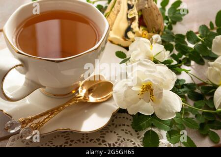 Tasse Tee und Zweig von kleinen weißen Rosen auf rustikalem Tisch, in weißen und goldenen Farben Stockfoto