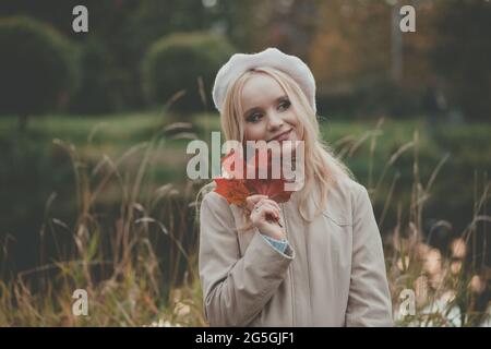 Outdoor-Porträt von jungen schönen glücklich lächelnden Frau mit langen Haaren trägt beige Baskenmütze und Mantel halten roten Herbstlaub im Herbst Park Stockfoto