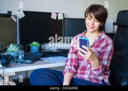 Lächelnde Frau, die auf den Telefonbildschirm blickt, während sie auf ihrem Heimbüro oder Arbeitsplatz im Büro sitzt. Machen Sie eine Pause. Lesen der guten Nachrichten. Remote-Arbeit Stockfoto