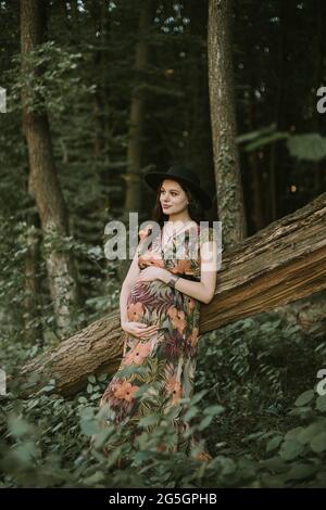 Schwangere in farbenfrohem Kleid und schwarzem Hut, posiert in einem grünen Sommerwald und steht in der Nähe des großen gefallenen Baumes Stockfoto