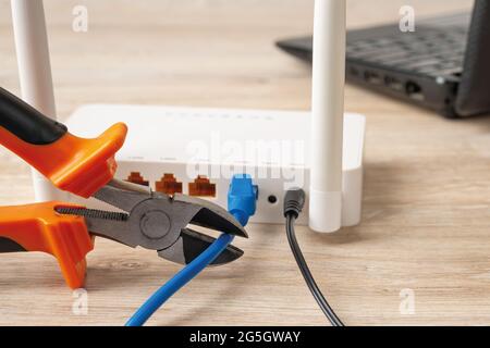 Nahaufnahme von Drahtabschneidern, die bereit sind, das Netzwerkkabel zu schneiden, das mit dem weißen wlan-Router auf einem Holzschreibtisch verbunden ist. Zugriff auf das Internet verbieten. Stockfoto