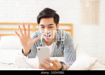 Hübscher junger Mann, der auf dem Bett liegt, ein Mobiltelefon benutzt, um einen Videoanruf zu tätigen und auf den Bildschirm winkt, um sich gegenseitig zu begrüßen Stockfoto