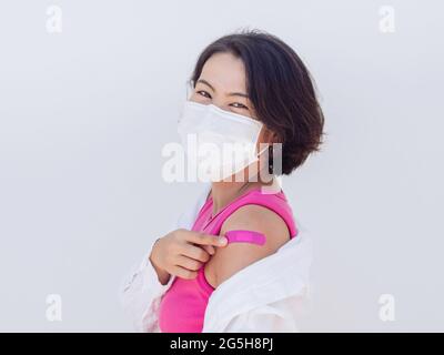 Impfungen, geimpfte Menschen Konzept. Glückliche asiatische Frauen tragen Gesichtsmaske und rosa ärmelloses Hemd zeigen Verband auf eigene Schulter mit Lächeln nach hinten Stockfoto