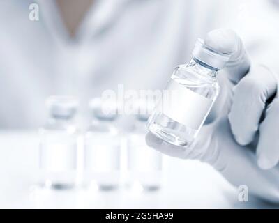 Medizinische Impfstofffläschchen mit weißem Rohling in der Hand des Wissenschaftlers mit weißen Gummihandschuhen. Nahaufnahme einer Hand, die Impfstoffglasflaschen für Injee hält Stockfoto