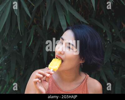 Frau, die Eis isst. Glücklich schöne asiatische Frau trägt beiläufige rosa Tank Top hält gelben Popsicle auf grünen tropischen Palmblättern Hintergrund, aus Stockfoto