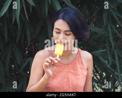 Frau, die Eis isst. Glücklich schöne asiatische Frau trägt beiläufige rosa Tank Top hält gelben Popsicle auf grünen tropischen Palmblättern Hintergrund, aus Stockfoto