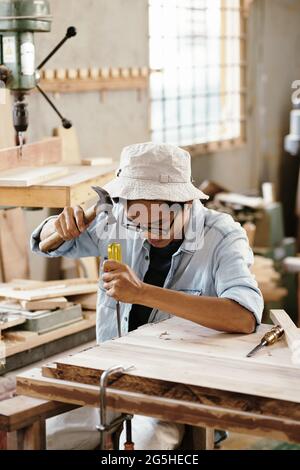 Seriös junger Zimmermann, der in der Werkstatt arbeitet und das Holz mit Meißel und Hammer schnitzt Stockfoto