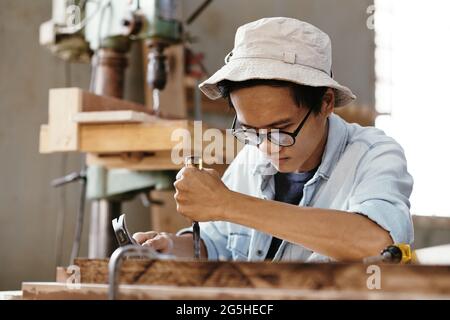 Konzentrierter kreativer junger Mann in den Gläsern, die Holz mit Meißel und Hammer in der Werkstatt schnitzen Stockfoto