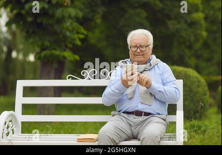 Glücklicher älterer Mann, der ein Mobiltelefon benutzt, während er auf einer Bank in einem grünen Sommerpark sitzt Stockfoto