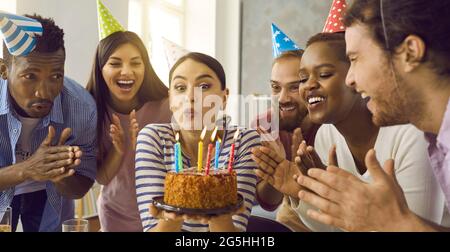 Freunde applaudieren, als das Geburtstagskind ein lustiges Gesicht macht und Kerzen auf ihren Kuchen bläst Stockfoto