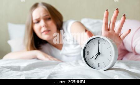 Verschlafenes junges Mädchen, das im Schlafanzug aufwacht, schaltet den Wecker eilig aus. Morgens Routine und spät vom Wecker im Bett aufwachen. Frau kommt spät Stockfoto