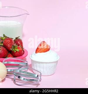 Cupcake mit Creme und einer halben Erdbeere, Schneebesen aus dem Mixer, Ei, Milch auf rosa Hintergrund. Selektiver Fokus. Stockfoto